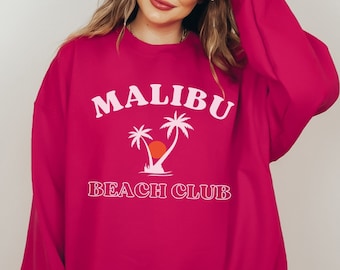 Op de afbeelding: Een roze sweatshirt met de tekst "MALIBU BEACH CLUB" in wit, met een palmboomgrafiek met een ondergaande zon.