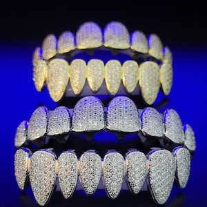 Può includere: Due set di grillz color oro e argento. Il set superiore è dorato, incrostato di piccole pietre scintillanti. Il set inferiore è argentato, anch'esso ricoperto di piccole pietre scintillanti. Entrambi i set sono esposti su uno sfondo blu scuro.