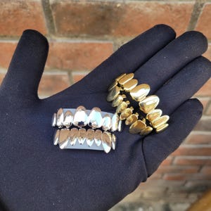 Può includere: Sono esposti due set di grillz per denti. Un set è color argento e l'altro è color oro. I grillz sono mostrati su una superficie nera. I grillz per denti sono un accessorio di moda.