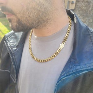 May include: A gold-colored chain necklace with a curb link design. The necklace has a rectangular clasp with small, clear stones. The necklace is worn over a gray t-shirt and a black leather jacket.