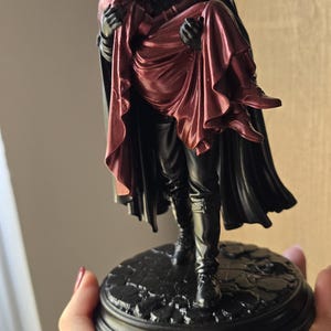May include: A black and metallic red figurine depicting a figure in a dark cloak carrying another figure draped in a deep red hooded garment. The sculpture stands on a circular, textured black base with a cobblestone pattern and smooth edges.