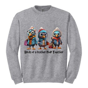 Puede incluir: Sudadera gris con tres pájaros de dibujos animados con gafas, sombreros y chaquetas, cada uno con bolsas de la compra. El texto "Birds of a feather SHOP Together" está impreso debajo.
