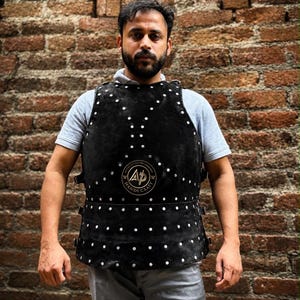 Può includere: Un gilet in pelle nera con borchie metalliche color argento disposte in file verticali e orizzontali. Sul petto è presente un logo circolare con la scritta AP HANDI CRAFT. Il gilet è dotato di fibbie laterali per la regolazione.