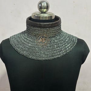 May include: A chainmail collar, displayed on a black mannequin. The collar is made of small, interlocking metal rings, with a leather trim at the neck. A metal emblem with the letters "AP" is visible on the front.