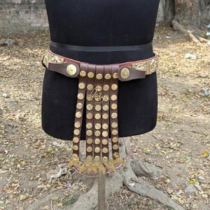 May include: A brown leather belt with gold-colored metal accents. The belt features a series of hanging leather strips adorned with gold studs and decorative elements. The belt is displayed on a black mannequin.