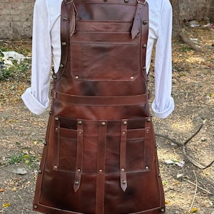 May include: A brown leather apron with multiple pockets and adjustable straps. The apron has brass-colored rivets and is displayed on a mannequin. The apron is designed for protection and utility.