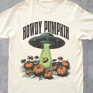 May include: Cream-coloured t-shirt with a graphic of a green UFO beaming a pumpkin into a pumpkin patch. The text "HOWDY PUMPKIN" is above the UFO. The shirt has short sleeves and a crew neck.