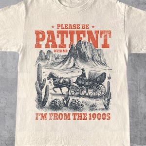 May include: Cream-colored t-shirt with a vintage-style graphic. The design features a horse-drawn carriage in a desert landscape, with text that reads "PLEASE BE PATIENT WITH ME I'M FROM THE 1900S".