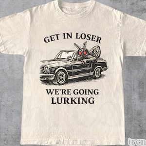 Puede incluir: Camiseta de color natural con un gráfico de una polilla conduciendo un coche clásico. El texto "GET IN LOSER WE'RE GOING LURKING" está impreso encima y debajo del coche. La camiseta tiene mangas cortas y cuello redondo.