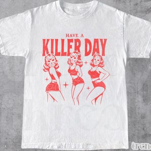 Könnte beinhalten: Weißes T-Shirt mit roter Grafik. Die Grafik zeigt den Text "HAVE A KILLER DAY" über drei Illustrationen von Frauen, die Hockeymasken und rote Outfits tragen. In der unteren linken Ecke des T-Shirts steht das Wort "WHITE".