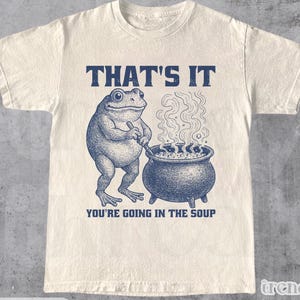 Pode incluir: Camiseta de cor natural com um gráfico azul marinho de um sapo mexendo em um caldeirão. O texto "THAT'S IT" e "YOU'RE GOING IN THE SOUP" estão acima e abaixo da ilustração do sapo.