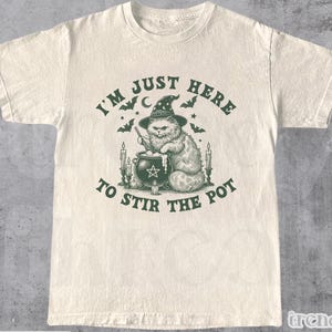 Puede incluir: Camiseta de color natural con un gráfico verde de un gato con sombrero de bruja revolviendo un caldero. El texto dice "I'M JUST HERE TO STIR THE POT". La camiseta tiene cuello redondo.