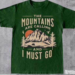 Puede incluir: Camiseta verde bosque con un gráfico de montaña. El diseño incluye una cordillera, un sol poniente, árboles y el texto "The Mountains Are Calling And I Must Go."