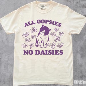 Puede incluir: Camiseta color crema con el texto morado "ALL OOPSIES NO DAISIES", un gráfico de gato y flores. La camiseta está etiquetada como "NATURAL" en la parte inferior izquierda y tiene la marca "trendy threads" en la parte inferior derecha.