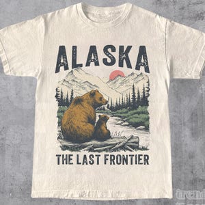 May include: Cream-coloured t-shirt featuring a graphic of two bears in a forest scene with mountains and the text "ALASKA" and "THE LAST FRONTIER". The shirt also has the word "NATURAL" printed at the bottom.