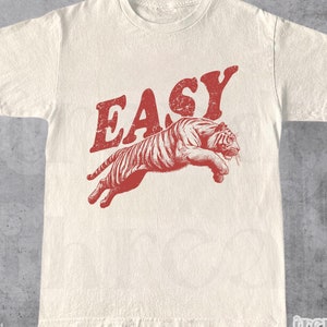 May include: Cream-colored t-shirt with a red graphic of a leaping tiger and the word "EASY" in a distressed font. The shirt is made of a soft, natural fabric. The brand name "trendy threads" is visible in the bottom right corner.