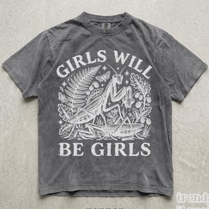 Op de afbeelding: Een antracietgrijs T-shirt met korte mouwen en een witte grafische print van een bidsprinkhaan omringd door varens, bessen en vlinders. De tekst GIRLS WILL BE GIRLS is in een vet, gebogen lettertype boven en onder de illustratie gedrukt.