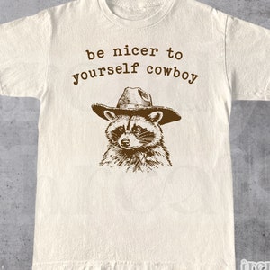 May include: A beige t-shirt with a brown raccoon wearing a cowboy hat and the text "be nicer to yourself cowboy".