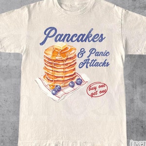 Könnte beinhalten: Ein weißes T-Shirt mit einem Aufdruck einer Stapel Pancakes mit Sirup und Blaubeeren. Der Text "Pancakes & Panic Attacks" befindet sich über den Pancakes und "buy one get one" darunter. Das Shirt ist in Naturfarbe.