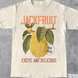 May include: A beige t-shirt featuring a vintage-style illustration of a jackfruit. The text "JACKFRUIT" is printed in orange above the fruit. Below the fruit is a circular sticker that says "100% FRESH" in orange text. The text "EXOTIC AND DELICIOUS" is printed in orange below the fruit.