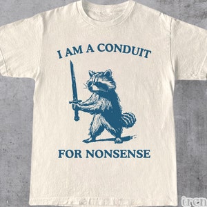 May include: A white t-shirt with a blue graphic of a raccoon holding a sword. The text "I am a conduit for nonsense" is printed below the raccoon.