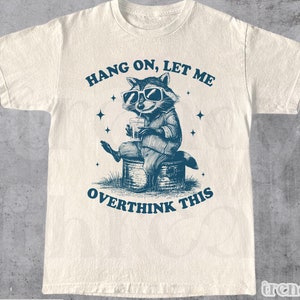 May include: A white t-shirt with a blue illustration of a raccoon wearing sunglasses and drinking from a can. The text "Hang on, let me overthink this" is printed on the shirt.