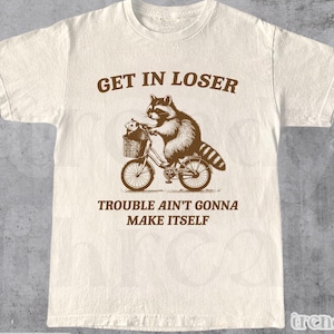 Könnte beinhalten: Ein beiges T-Shirt mit einer braunen Grafik eines Waschbären, der ein Fahrrad fährt, mit einem kleineren Waschbären im Korb. Der Text "GET IN LOSER" befindet sich über der Grafik und "TROUBLE AIN'T GONNA MAKE ITSELF" unter der Grafik.