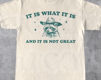 Peut inclure: Un t-shirt beige avec un graphique vert d'un raton laveur portant un chapeau de cowboy. Le texte "IT IS WHAT IT IS AND IT IS NOT GREAT" est imprimé sur le t-shirt.