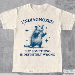 Op de afbeelding: Crèmekleurig T-shirt met een blauwe illustratie van een opossum en de tekst "UNDIAGNOSED BUT SOMETHING IS DEFINITELY WRONG." Het shirt heeft een ronde hals en korte mouwen. Het woord "NATURAL" staat onderaan.
