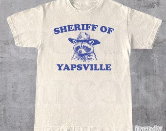 Puede incluir: Una camiseta blanca con un gráfico azul de un mapache con un sombrero de sheriff. El texto "SHERIFF OF YAPSVILLE" está impreso en azul debajo del mapache.