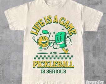 Pode incluir: Uma camiseta branca com um design gráfico verde e amarelo. O gráfico apresenta dois personagens de desenho animado jogando pickleball. O texto "Life is a game and pickleball is serious" está impresso na camiseta.