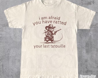 Op de afbeelding: Een wit T-shirt met een bruine illustratie van een rat die een cowboyhoed draagt en een pistool vasthoudt. De tekst "i am afraid you have ratted your last tatouille" is op het T-shirt gedrukt.