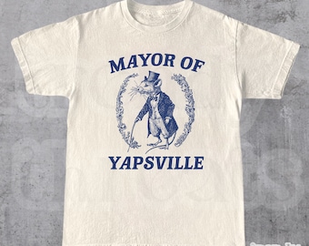 May include: A white t-shirt featuring a blue graphic of a mouse wearing a top hat and holding a cane. The text "MAYOR OF YAPSVILLE" is printed below the graphic.