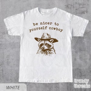 May include: White t-shirt with a brown illustration of a raccoon wearing a cowboy hat. The text "be nicer to yourself cowboy" is printed above the raccoon.