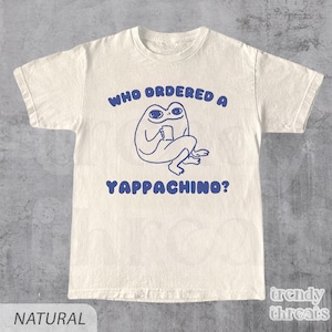 May include: A cream-coloured t-shirt featuring a blue illustration of a frog with a cup, and the text "WHO ORDERED A YAPPACHINO?" in blue. The shirt has a crew neck and short sleeves.