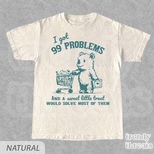 Op de afbeelding: Een wit T-shirt met een turquoise afbeelding van een beer die een winkelwagen vol boodschappen duwt. De tekst "Ik heb 99 problemen en een zoete kleine traktatie zou de meeste oplossen" staat onder de beer.