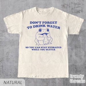 K&ouml;nnte beinhalten: Ein wei&szlig;es T-Shirt mit einem blauen Frosch-Motiv, das eine Wasserflasche h&auml;lt. Der Text auf dem Shirt lautet "Vergiss nicht, Wasser zu trinken, damit du hydriert bleibst, w&auml;hrend du leidest."