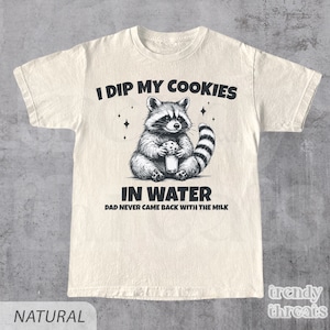 May include: A beige t-shirt with a black and white illustration of a raccoon holding a cookie and a glass of water. The text "I dip my cookies in water. Dad never came back with the milk." is printed on the shirt.