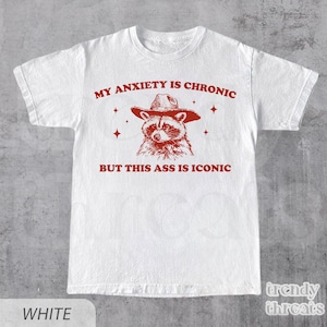 Puede incluir: Camiseta blanca con un gráfico rojo de un mapache con sombrero de vaquero. El texto "MY ANXIETY IS CHRONIC BUT THIS ASS IS ICONIC" está impreso en rojo. La camiseta tiene cuello redondo y mangas cortas.