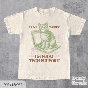 Può includere: Una t-shirt beige con un gatto verde seduto su un laptop. Il testo sulla t-shirt recita "Don't Worry I'm From Tech Support".