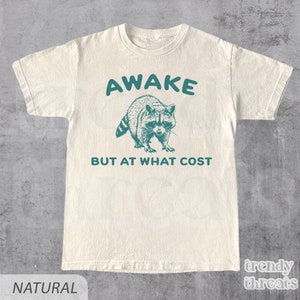May include: A white t-shirt with a teal green graphic of a raccoon and the text "AWAKE BUT AT WHAT COST".