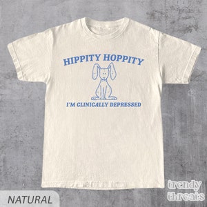 Może przedstawiać: Beżowa koszulka z niebieskim nadrukiem przedstawiającym królika z tekstem "Hippity Hoppity I'm Clinically Depressed".