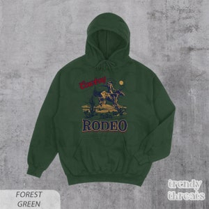 May include: Forest green hooded sweatshirt with a vintage-style illustration of a cowboy riding a horse. The text "Cowboy Rodeo" is printed in red and gold on the front of the sweatshirt.