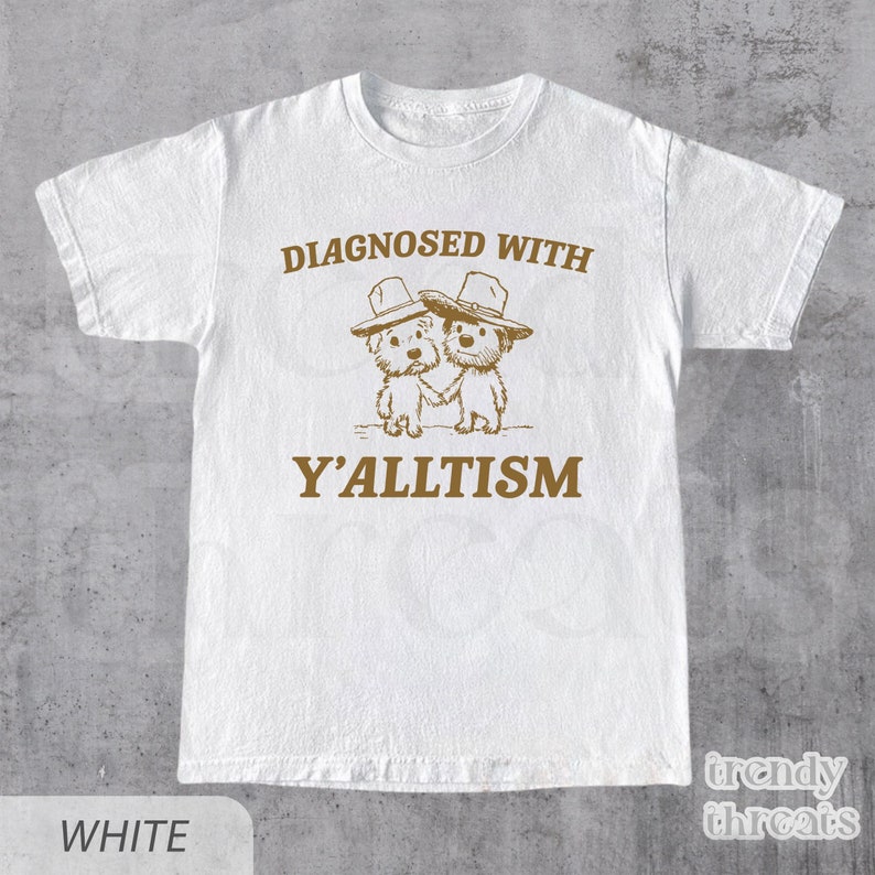 May include: White t-shirt with a humorous graphic. The design features two cartoon dogs wearing hats, holding hands, and the text "DIAGNOSED WITH Y'ALLTISM" in brown. The shirt has a crew neck and short sleeves. The brand name "trendy threats" is in the bottom right corner.