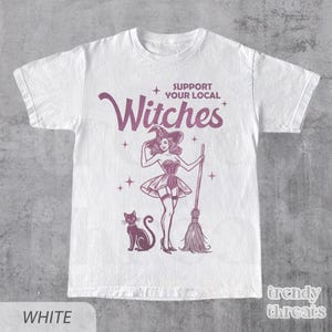 Flat lay white color shirt with quote 'Support Your Local Witches' and retro Halloween witch graphic.
