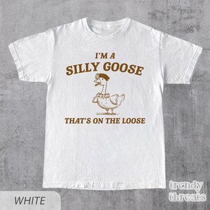 Op de afbeelding: Wit T-shirt met een bruine illustratie van een gans met een hoed en de tekst "I'm a Silly Goose That's on the Loose."