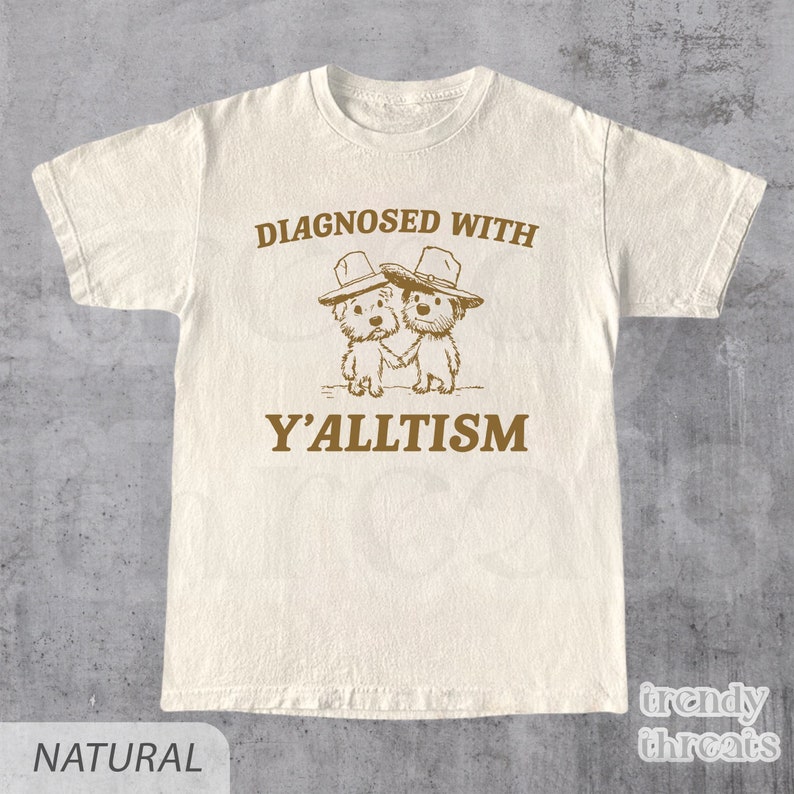 May include: Natural-colored t-shirt with the text "DIAGNOSED WITH Y'ALLTISM" in brown. The graphic features two cartoon dogs wearing hats and holding hands. The shirt has a crew neck and short sleeves.