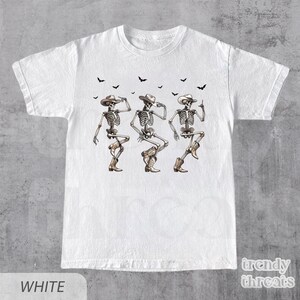 May include: White t-shirt with a graphic of three skeletons wearing cowboy hats and boots, dancing. The skeletons are outlined in black and have a light brown color.