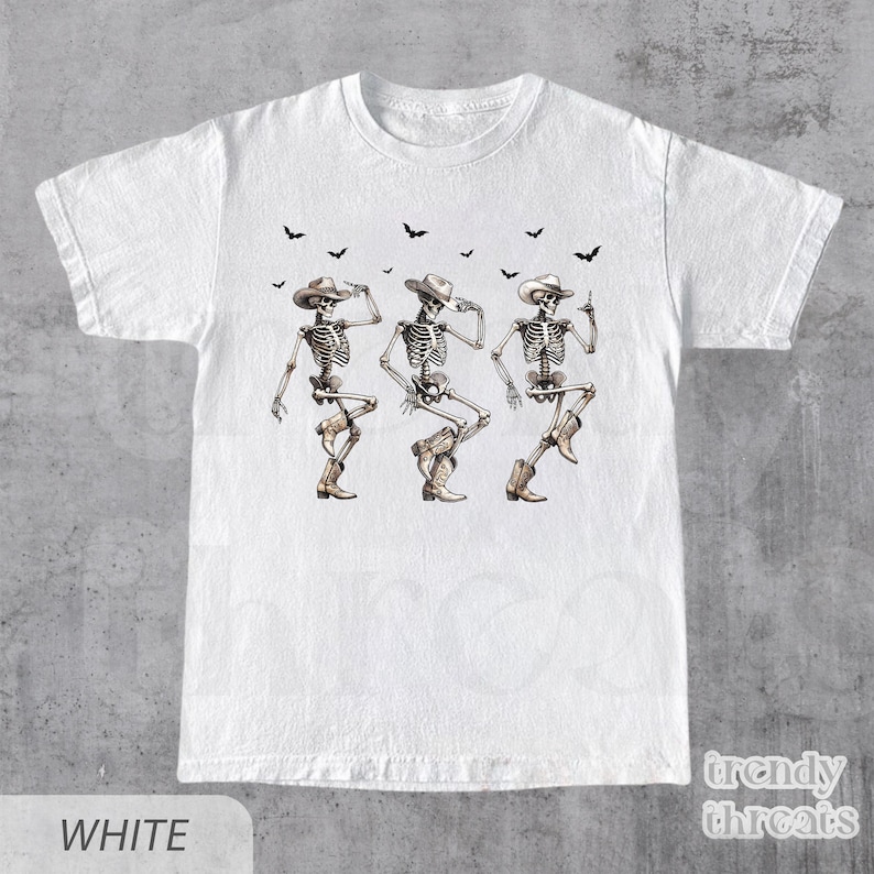 May include: White t-shirt with a graphic of three skeletons wearing cowboy hats and boots, dancing. The skeletons are outlined in black and have a light brown color.