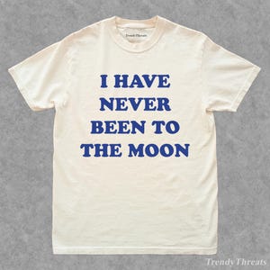 Puede incluir: Camiseta color crema con el texto azul "I HAVE NEVER BEEN TO THE MOON". La camiseta tiene cuello redondo y mangas cortas. El nombre de la marca "Trendy Threads" está impreso en el cuello.